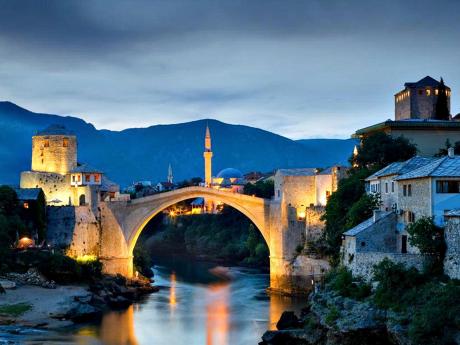 Večerní Mostar s osvětleným mostem, strážní věží a minaretem v pozadí